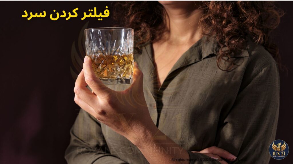 تصویر نزدیک از لیوان ویسکی شفاف که نتیجه‌ی فیلتر کردن سرد است، در دست فردی با نور ملایم و پس‌زمینه‌ی تیره | Lux21